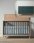 Wooden crib with green and white bedding next to a matching dresser in a minimalistic room.