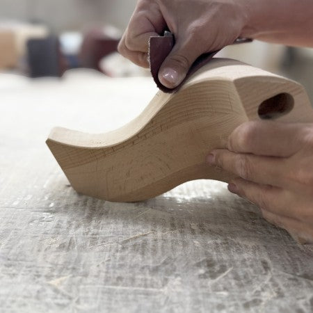 Person sanding a wooden shoe last with a sanding block on a textured surface.